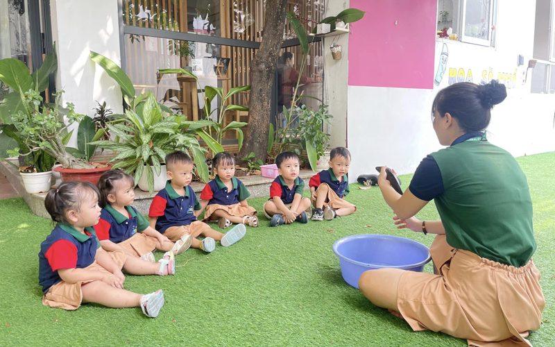 Các bé ngồi trên thảm cỏ ngoài sân nghe cô hướng dẫn